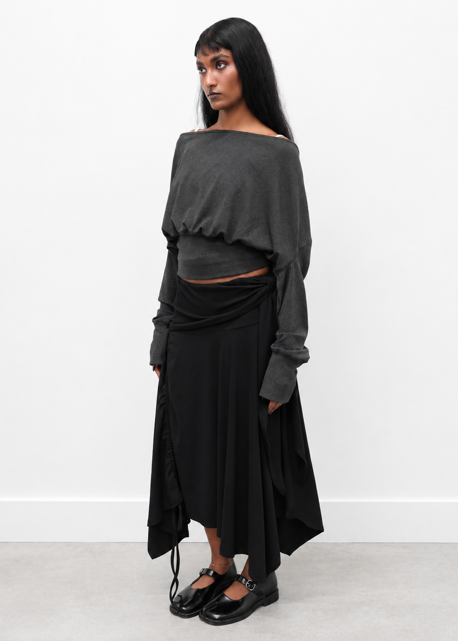 A model stands in a minimalist studio setting wearing a dark gray off-the-shoulder cropped long-sleeve top and a black draped midi skirt, paired with black Mary Jane shoes.