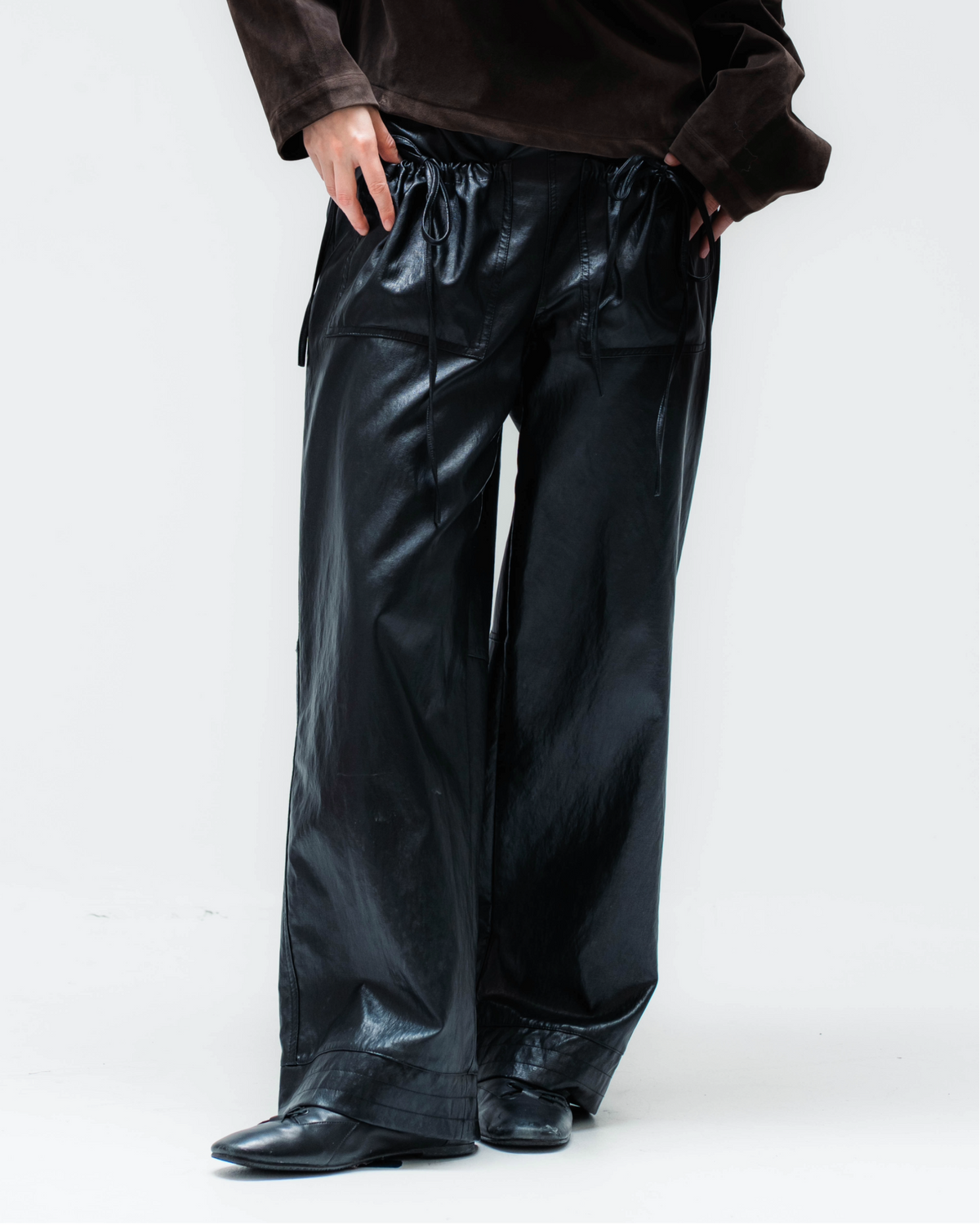 A model wearing a dark brown off-the-shoulder velvet top and shiny black wide-leg pants, standing against a plain white studio background.