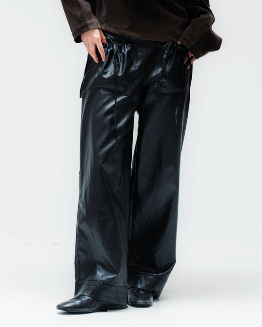 A model wearing a dark brown off-the-shoulder velvet top and shiny black wide-leg pants, standing against a plain white studio background.